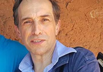 La foto muestra a Julien Harneis, inclinado hacia el hombro de otra persona. Lleva una chaqueta de mezclilla, una camisa azul y se para frente a una pared de cemento color barro.