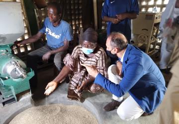 El Coordinador Residente, Vincent Martin, y una líder comunitaria (ambos con mascarilla) arrodillados cerca de una máquina, mientras la líder ejemplifica cómo se llena y se procesas los granos. En el fondo hay dos hombres con vestimenta de operario y de seguridad.