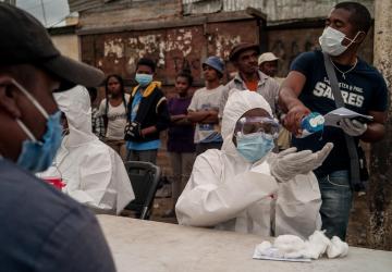 Image shows people waiting behind a table of healthcare workers wearing head-to-toe personal protective equipment. One man standing behind the healthcare workers pours had sanitizer onto the hands of one of the workers, while a man sits in front of her and waits.