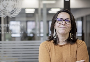 Una mujer con una camisa naranja y gafas negras se encuentra en una oficina de las Naciones Unidas con los brazos cruzados.
