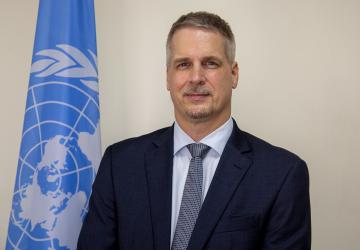 A UN official in a suit and tie poses for a photo next to a blue UN flag