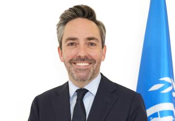 A man in a black suit and tie stands in front of a blue UN flag
