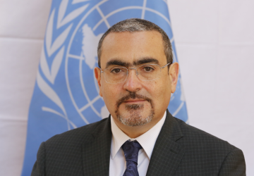 A UN official in a suit and tie poses for a photo in front of a blue UN flag