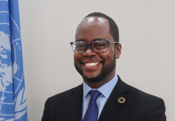 A UN official in a suit and tie smiles and stands next to a UN flag 