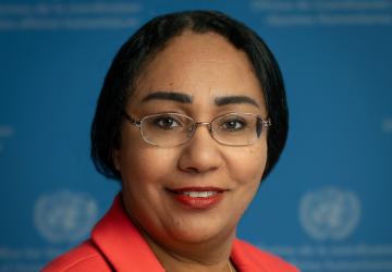 A woman in an orange coloured suit wearing glasses standing in front of a blue background with the UN logo