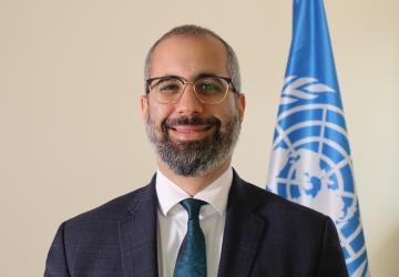 A man in a black suit stands in front of a blue UN flag