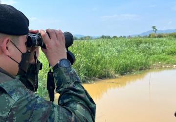 A man wearing a mask, dressed in camo-military garb and a black beret, looks out at a murky river with binoculars.