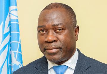 A man in a dark grey suit and blue tie standing next to a blue UN flag.