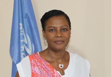 A woman in a white and pink shirt stands in front of a blue UN flag.