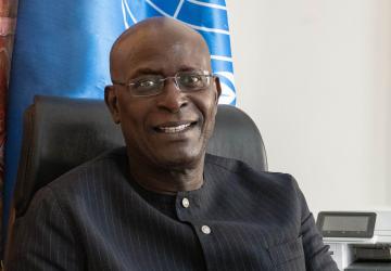 A man in a black suit and glasses sits in a chair with a blue UN flag behind him.