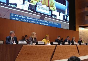 A long podium that is seating seven people, men and women, in various colored clothes. Behind them is a giant screen with a projection of a woman in a yellow dress, the speaker, Amina Mohammed.