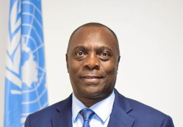 A man in a blue suit and blue tie stands in front of a blue UN flag