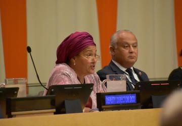 A woman in a red headscarf and pink shirt speaks into a microphone as a man in a black suit listens on