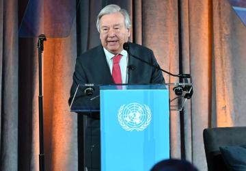 A man in a black suit, the UN Secretary-General speaks into a microphone at a podium with the blue UN flag hanging over it