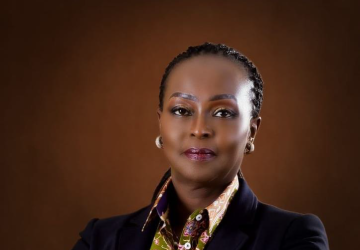 A woman in a black suit and golden brown scarf with her arms crossed against a brown background