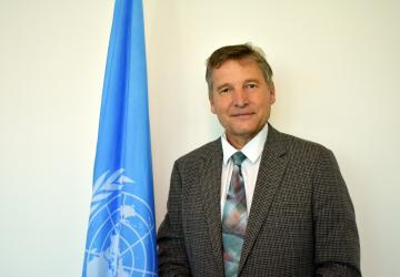 A man in a dark suit and a multicoloured tie stands next to a blue flag with the UN logo on it