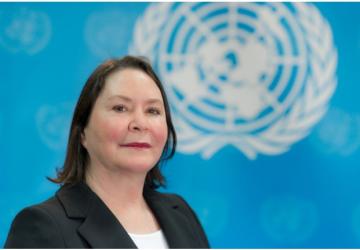 A woman in a black suit and white shirt stands in front of a blue flag with the UN logo on it