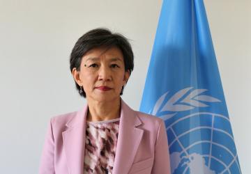 A woman in a pink blazer and shirt stands in front of a blue UN flag