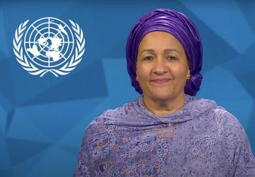 A woman in a purple dress and headdress stands in front of a blue screen with a white UN logo