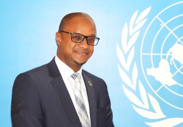 A man in a charcoal grey suit with a grey tie and wearing spectacles stands in front of blue wall with a white UN logo