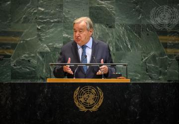 A man, the Secretary-General of the United Nations, in a dark suit and tie, gesticulates as he addresses people from a podium