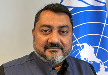 A man in a dark suit stands in front of a blue flag with the UN logo on it.