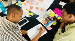 شابان يجلسان على مكتب عليه الكثير من الأوراق ولوحة ملصقات.