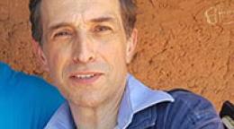 Photo shows Julien leaning towards the shoulder of another person. He's wearing a denim jacket, blue shirt and stands in front of a mud-coloured cement wall. 