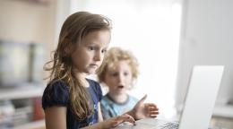 A little girl is shown using a laptop, while her younger brother stands in awe. 