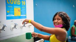 Une jeune Vénézuélienne vêtue d’un débardeur jaune et portant un masque rose descendu au niveau du menton se tient debout dans une station d’eau potable. Elle regarde la caméra en arborant un grand sourire tandis qu'elle se prépare à remplir d’eau une tasse de couleur verte.