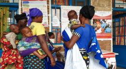 Trois femmes portant chacune un bébé se tiennent devant la porte d'un centre médical et se renseignent auprès d’un membre féminin du personnel assis à une table à l’extérieur du centre. Plusieurs affiches contenant des informations sur la COVID-19 et sur la vaccination contre d’autres maladies infectieuses ont été collées sur le mur externe du centre.
