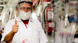 Un marchand portant une visière de protection et une blouse blanche se tient près d’un étal de produits alimentaires protégé par des bâches en plastique. Il regarde la caméra en levant le pouce.