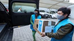 Dans un parking, un homme et une femme membres du personnel de l'ONU s'apprêtent à déposer un grand carton de fournitures offertes par le PNUD dans le coffre d'un véhicule. Chacun d'eux porte un masque de protection respiratoire.