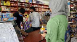 Une femme portant des vêtements clairs paye ses achats dans un supermarché à l’aide d’un bon d’achat électronique. Sur le comptoir se trouve un flacon de gel hydroalcoolique. Derrière le comptoir, un homme portant un polo noir et un masque de protection finalise la procédure de paiement. En arrière-plan, on aperçoit un autre homme et plusieurs rayons de produits alimentaires.