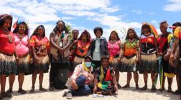 La delegación de ONU Mujeres y las mujeres miembros del Consejo Provincial de Hela están reunidas para la inauguración de la oficina de la ONU en Tari, provincia de Hela. La mayoría de las mujeres están de pie frente a la cámara, mientras que otras dos mujeres están sentadas en el suelo en primer plano. Muchas mujeres del consejo provincial visten ropas tradicionales de colores vivos.