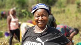 Une agricultrice adresse un sourire enjoué à la caméra, tandis que d'autres femmes procèdent à la récolte en arrière-plan.