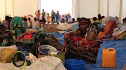 Several people sitting in an indoors market with baskets and a gas can.