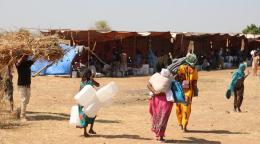 Deux femmes et une fille marchent en direction d’un camp, dos à la caméra, en transportant des bidons, des matelas et des couvertures. En arrière-plan, on aperçoit de nombreuses personnes s’abritant sous des tentes.