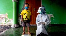 Un niño está parado sobre una balanza. Lleva una máscara protectora y mira a la cámara. A su izquierda, un cuidador que usa equipo de protección personal de pies a cabeza y una visera protectora se arrodilla a su lado y lo mira con una sonrisa.