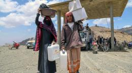 Un homme et une femme marchent côte à côte en direction de la caméra en transportant divers objets. Ils viennent de recevoir un kit distribué par l’ONU dans le cadre du Mécanisme d’intervention rapide. En arrière-plan, on aperçoit plusieurs personnes et plusieurs motos et véhicules stationnés sous une sorte de préau au milieu d’un terrain désertique.