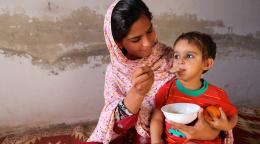 A young woman holds a child in her lap while she feeds him from a bowl.