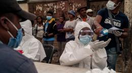 Image shows people waiting behind a table of healthcare workers wearing head-to-toe personal protective equipment. One man standing behind the healthcare workers pours had sanitizer onto the hands of one of the workers, while a man sits in front of her and waits.
