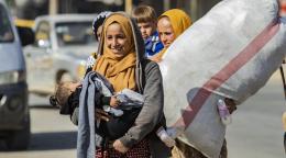 A woman smiles as she carries a sleeping child. Another woman walks behind her as she carries a large bag. 