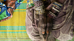 A woman's hands, with several bracelets, rest on her knees on top of a bright colored cloth.