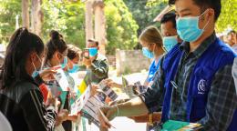 Several members of the IOM staff in blue vests and blue facemasks hand out informational pamphlets to a group of women. 