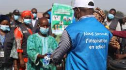 Un voluntario de las Naciones Unidas con un chaleco azul le entrega una bolsa a un hombre. Varias personas están detrás del hombre que recibe el paquete.