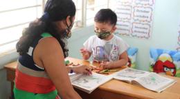 A woman and a child in face masks look over coloring books on a wooden table. 