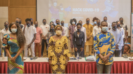 A large group of people in face masks stand facing the camera in front of projector displaying the words "Hack COVID-19."