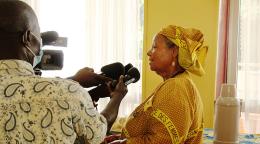 A woman in a gold dress and headscarf speaks into a microphone held by a man in a white shirt with black designs. 