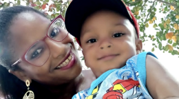 Gros plan sur une femme portant des lunettes, souriant à la caméra et tenant un jeune enfant dans les bras. L’enfant porte une casquette et sourit lui aussi à la caméra.